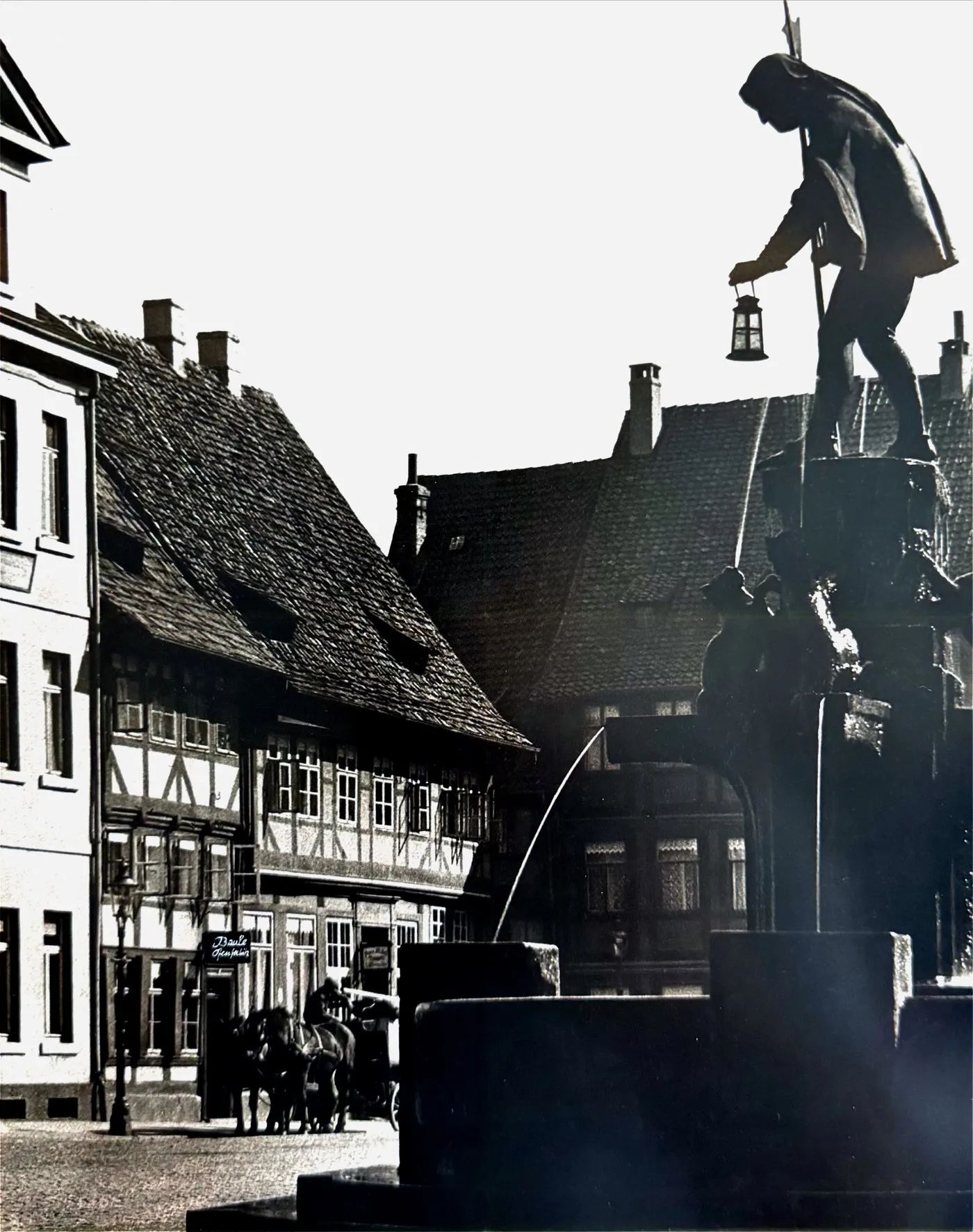 Historische Aufnahme vom Neustädter Markt in Hildesheim: der Marktbrunnen mit der Figur des Nachtwächters, dahinter die Fachwerkhäuser mit dem Geschäft von Ofen Baule
